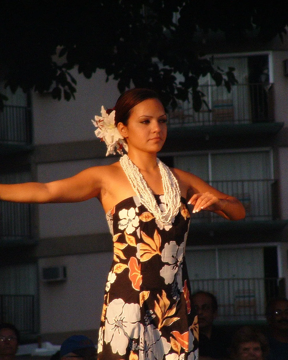 A hula dancer in a floral dress and white flower lei performs with her arms outstretched at golden hour